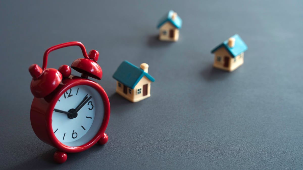 Red alarm clock in foreground with houses blurred behind, representing urgent timelines to sell